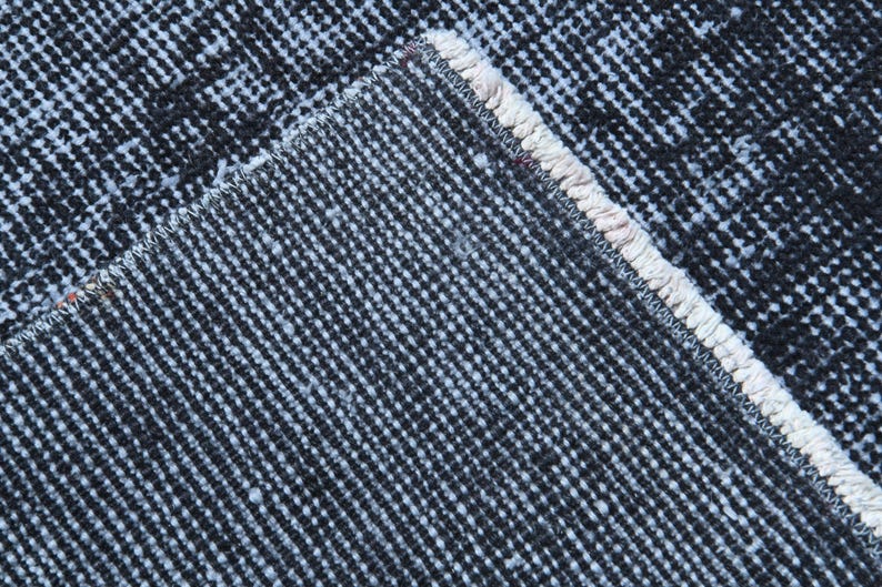 May include: A close-up view of the underside of a rug, showing the dark charcoal gray woven backing material with a distinct grid-like texture and a thick, light cream-colored stitched edge binding that runs along the diagonal corner seam.