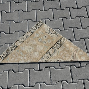 May include: A triangular beige rug with a geometric pattern in brown and blue. The rug is lying on a gray concrete surface.