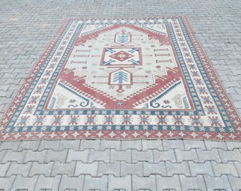 Könnte beinhalten: Rechteckiger Teppich mit einem zentralen geometrischen Design in Rot, Creme und Blau. Der Teppich hat einen Rand mit sich wiederholenden Mustern und liegt auf einer grauen Ziegeloberfläche. Das Design des Teppichs umfasst stilisierte Baum- und Pfeilmotive.