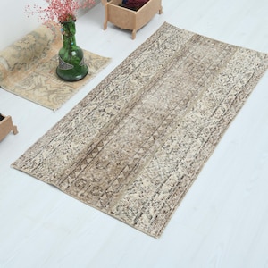 Puede incluir: Una alfombra rectangular beige y marrón estampada con un aspecto envejecido y vintage. La alfombra está sobre una superficie blanca, con una alfombra enrollada y un jarrón de cristal verde con flores rosas en el fondo. También se ve una pequeña caja de madera con bolas de hilo de colores.