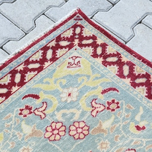 May include: A vintage rug with a light blue background and a red and white border. The rug has a floral pattern with yellow, pink, and white flowers.