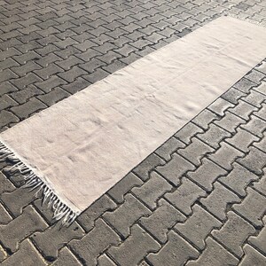 Könnte beinhalten: Ein weißer rechteckiger Teppich mit Fransen an den Rändern. Der Teppich liegt auf einer grauen Betonfläche.