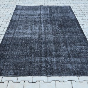 May include: A rectangular area rug featuring a distressed, faded black and dark charcoal gray color palette. The surface displays a worn, mottled texture with subtle variations in tone across the entire field, creating a weathered appearance.