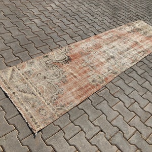 May include: A long, rectangular rug with a faded, distressed look. The rug has a light brown background with a pattern of faded, dark brown and black geometric shapes. The rug is fringed on all sides.
