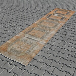 K&ouml;nnte beinhalten: Ein langer, rechteckiger Teppich mit einem verblassten orangefarbenen und grauen geometrischen Muster. Der Teppich liegt auf einer grauen Betonfl&auml;che.