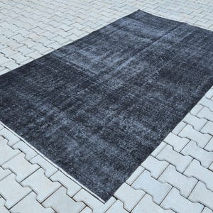 May include: A rectangular area rug featuring a distressed, faded black and dark charcoal gray color palette. The surface displays a worn, mottled texture with subtle variations in tone across the entire fabric, creating a weathered appearance.