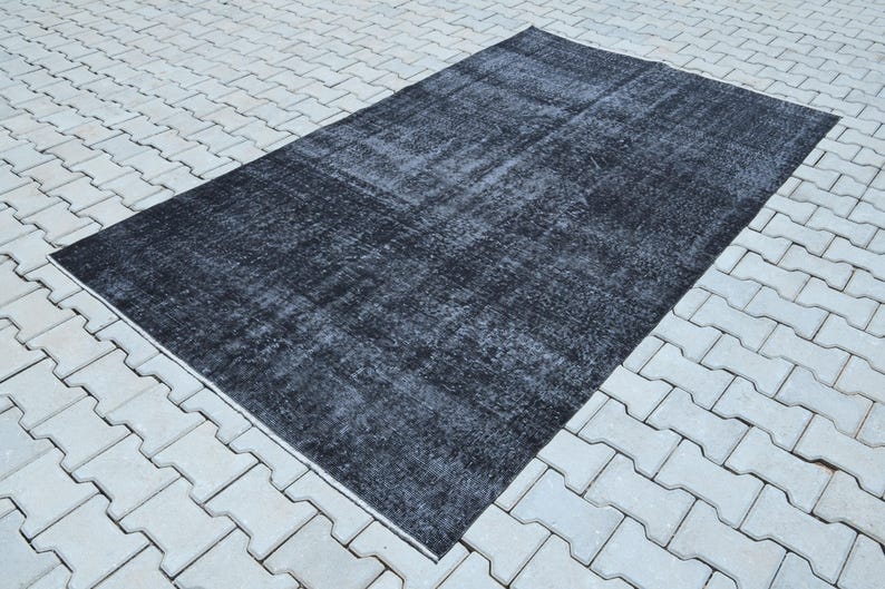 May include: A rectangular area rug featuring a distressed, faded black and dark charcoal gray color palette. The surface displays a worn, mottled texture with subtle variations in tone across the entire fabric, creating a weathered appearance.