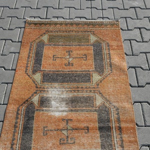 K&ouml;nnte beinhalten: Ein Vintage-Teppich mit einem geometrischen Muster in Orange-, Schwarz- und Wei&szlig;t&ouml;nen. Der Teppich hat einen abgenutzten und verblassten Look, der ihm einen rustikalen Charme verleiht.