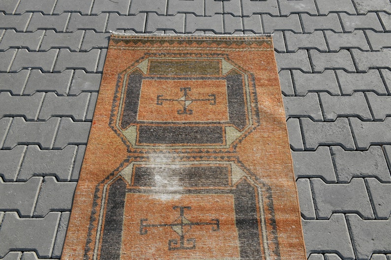 K&ouml;nnte beinhalten: Ein Vintage-Teppich mit einem geometrischen Muster in Orange-, Schwarz- und Wei&szlig;t&ouml;nen. Der Teppich hat einen abgenutzten und verblassten Look, der ihm einen rustikalen Charme verleiht.