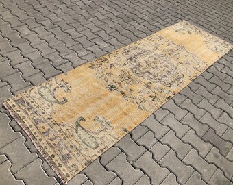 May include: A vintage runner rug with a faded floral pattern in shades of beige, brown, and green. The rug has a fringed edge and is laid out on a gray concrete surface.