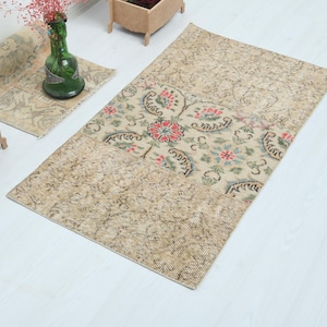 Könnte beinhalten: Ein rechteckiger, beigefarbener Teppich mit einem zentralen Blumenmuster in Rot, Grün und Blau. Der Teppich hat ein abgenutztes, Vintage-Aussehen. Ein aufgerollter Teppich und eine kleine Kiste mit bunter Wolle sind ebenfalls zu sehen.