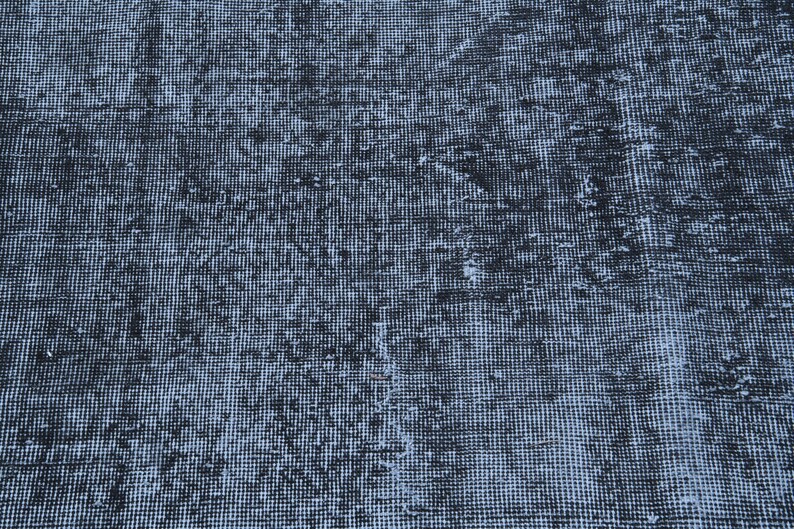 May include: A woven textile rug featuring a distressed, mottled pattern in shades of deep black and faded charcoal gray. The surface displays a dense, uniform grid of small, interconnected fibers with visible areas of lighter, worn-out texture.