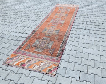 May include: A long, narrow rug with a geometric pattern in shades of orange, pink, and gray. The rug is laid out on a gray concrete surface.