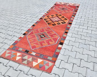 May include: A long, rectangular red and orange rug with a geometric pattern. The rug has a fringe border and is made of wool.