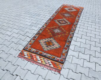 Puede incluir: Una alfombra larga y estrecha, de color naranja y marrón, con un patrón geométrico. La alfombra tiene flecos en ambos extremos.