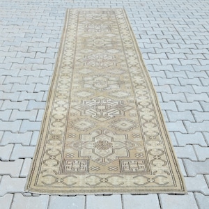 May include: A long, narrow runner rug featuring a muted beige and tan color palette. The rug displays a repeating geometric pattern with central diamond-shaped medallions and intricate border details in darker brown and cream tones.