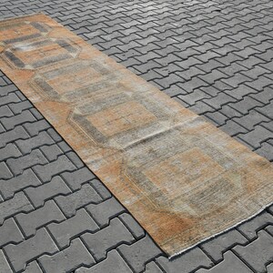 K&ouml;nnte beinhalten: Ein langer, rechteckiger Teppich mit einem geometrischen Muster in Beige-, Braun- und Graut&ouml;nen. Der Teppich liegt auf einer grauen Betonfl&auml;che.