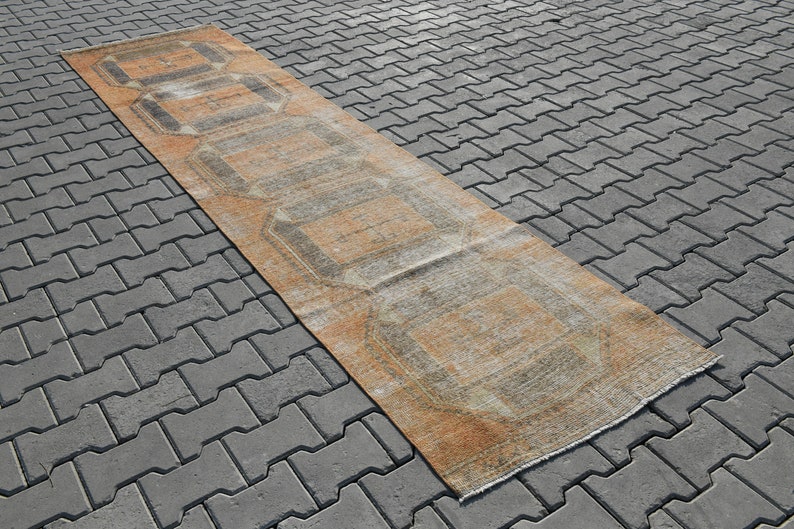 K&ouml;nnte beinhalten: Ein langer, rechteckiger Teppich mit einem geometrischen Muster in Beige-, Braun- und Graut&ouml;nen. Der Teppich liegt auf einer grauen Betonfl&auml;che.