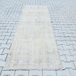Può includere: Un tappeto rettangolare beige chiaro dall'aspetto sbiadito. Il tappeto ha un design semplice e una frangia lungo un bordo. Il tappeto è posizionato su una superficie grigia a motivi.
