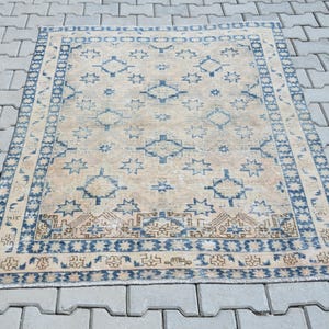 Puede incluir: Una alfombra rectangular con fondo beige y un patrón geométrico repetitivo de estrellas azules. La alfombra tiene un borde ancho azul con diseños lineales intrincados y flecos en los extremos, mostrando una apariencia desgastada y descolorida.