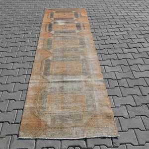 K&ouml;nnte beinhalten: Ein langer, rechteckiger Teppich mit einem verblassten orange-braunen geometrischen Muster. Der Teppich hat einen abgenutzten Look und ist aus einem weichen, gewebten Material gefertigt.