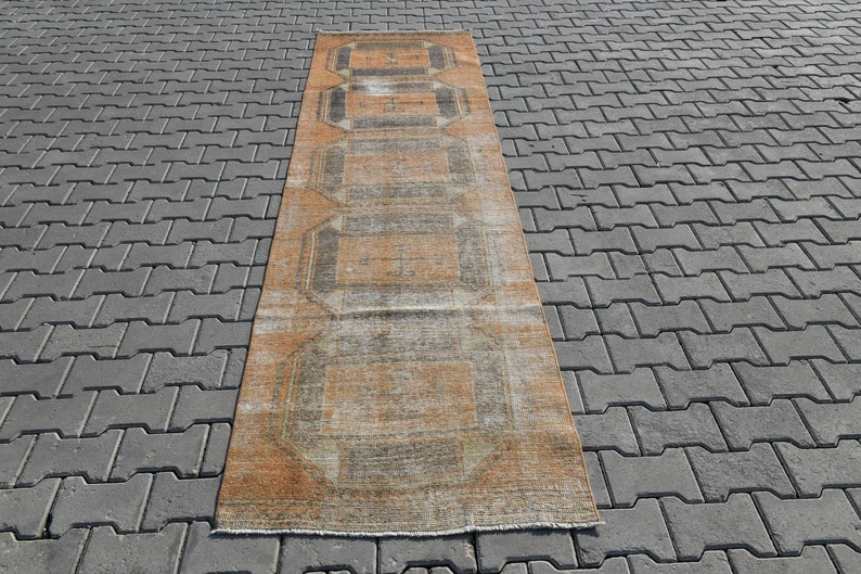 K&ouml;nnte beinhalten: Ein langer, rechteckiger Teppich mit einem verblassten orange-braunen geometrischen Muster. Der Teppich hat einen abgenutzten Look und ist aus einem weichen, gewebten Material gefertigt.