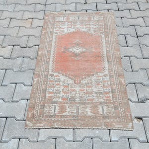 Puede incluir: Alfombra rectangular de estilo vintage con un diseño de medallón central en tonos coral y crema. La alfombra presenta intrincados bordes en tonos marrones y beige. Se encuentra sobre una superficie gris texturizada. Mide aprox. 120 cm x 60 cm.