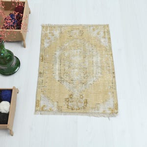 May include: A rectangular, beige rug with a distressed, vintage appearance. The rug features a central medallion design and fringe along the bottom edge. The rug is placed on a white surface, with decorative items nearby.