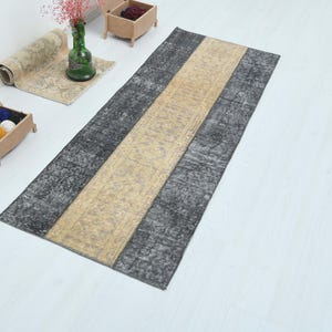 Può includere: Un tappeto rettangolare con una striscia centrale beige affiancata da sezioni grigio scuro. Il tappeto ha un aspetto invecchiato e vintage. Il tappeto è posto su una superficie bianca.