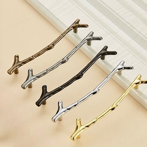 Może przedstawiać: Pięć różnych kolorowych gałek do szafek w kształcie gałęzi. Gałki są wykonane z metalu i mają rustykalny wygląd. Kolory to złoty, srebrny, czarny i dwa odcienie brązu.