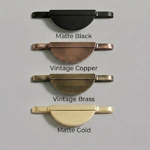 Op de afbeelding: Vier kastgrepen in matzwart, vintage koper, vintage messing en mat goud. Elke greep heeft een halfrond ontwerp met aan elke kant een rechthoekige handgreep. De tekst "Matte Black", "Vintage Copper", "Vintage Brass" en "Matte Gold" staat eronder.