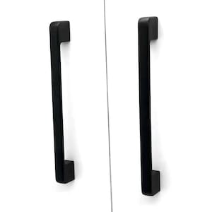 May include: Two vertical matte black cabinet handles with a rectangular profile and squared edges. Each handle features a long, straight bar design with two mounting points at the top and bottom, attached to a plain white surface.
