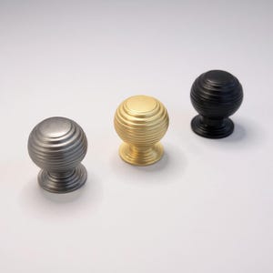 May include: Three decorative cabinet knobs in silver, gold, and black. Each knob has a spherical shape with horizontal ridged detailing. The knobs are designed with a base for mounting on furniture.