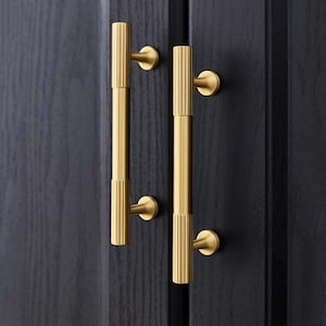 Puede incluir: Primer plano de dos manijas de puerta doradas. Cada manija tiene un diseño cilíndrico con ranuras verticales y una base circular. Las manijas están montadas en una puerta de madera oscura, creando un contraste de color y textura.