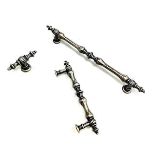 May include: Three antique silver-toned cabinet door pulls. Two are long and slender with ornate details. The third is a smaller, more traditional knob.