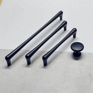 Peut inclure: Un ensemble de quatre pièces de quincaillerie d'armoire noir mat. L'ensemble comprend deux longues barres de tirage, une barre de tirage moyenne et un bouton rond. La quincaillerie est présentée sur une surface grise clair et blanche.