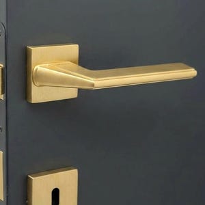 May include: A gold-colored door handle and keyhole plate set against a dark gray door. The handle has a rectangular base and a straight, slightly angled lever. The keyhole plate is square with a centered keyhole.