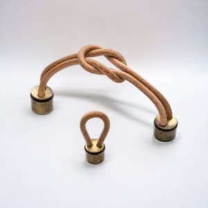 Puede incluir: Dos asas de cordón de cuero con tapas de latón. El asa más grande tiene un diseño anudado. El asa más pequeña es un simple bucle.