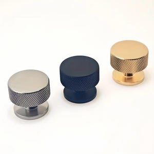 Puede incluir: Tres pomos de armario en diferentes colores: plateado, negro y dorado. Cada pomo tiene una parte superior redonda y texturizada y una base redonda más pequeña.