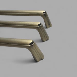 May include: Three antique brass cabinet pulls with a curved design. The pulls have a rectangular bar with a curved handle and a decorative base. The finish has a slightly distressed look.