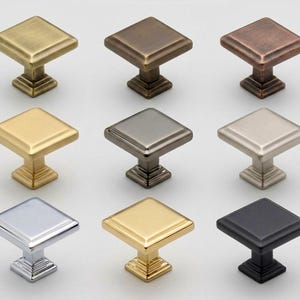 May include: Nine square cabinet knobs in various metallic finishes, including gold, bronze, silver, and black. Each knob features a square top and a decorative base, suitable for furniture or cabinetry.