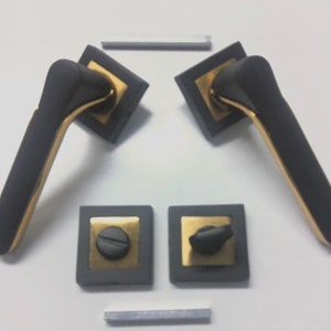 Op de afbeelding: Twee zwarte en gouden deurgrepen met vierkante rozetten. De grepen hebben een modern ontwerp met een gebogen vorm. De rozetten hebben een gouden rand.