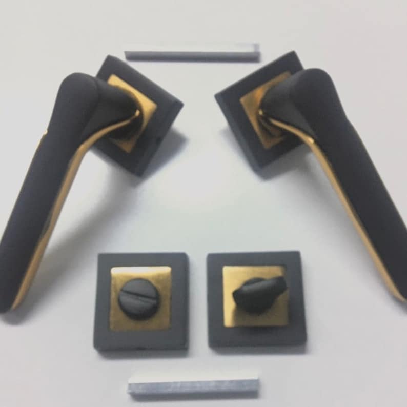 Op de afbeelding: Twee zwarte en gouden deurgrepen met vierkante rozetten. De grepen hebben een modern ontwerp met een gebogen vorm. De rozetten hebben een gouden rand.