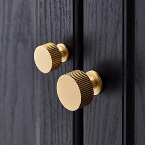 Op de afbeelding: Twee ronde gouden kastknoppen met een geribbeld ontwerp. De knoppen zijn gemonteerd op een zwart oppervlak.