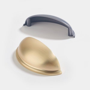 Puede incluir: Dos tiradores de armario, uno en oro cepillado y otro en acabado gris oscuro. Ambos tiradores tienen una forma curva en forma de taza.