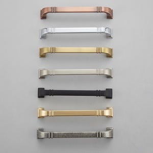 May include: A collection of seven horizontal cabinet pulls arranged in a vertical column. Each handle features a straight bar design with two vertical ridges near the ends and square mounting bases. The finishes include copper, chrome, brass, nickel, matte black, gold, and antique silver.