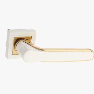 May include: A door handle featuring a white rectangular base plate and a horizontal lever. The lever has a white front surface framed by a polished gold-tone metal edge. The connection point between the base and the lever is also gold-tone.