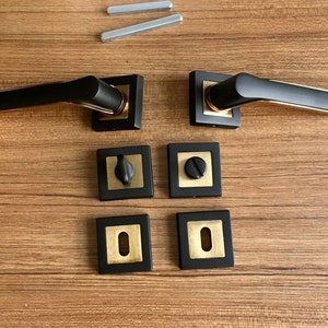 Op de afbeelding: Set zwarte deurgrepen met gouden accenten. De grepen zijn vierkant en hebben een modern ontwerp. De set bevat twee grepen, twee vierkante platen en twee ronde platen.
