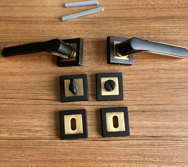 Op de afbeelding: Set zwarte deurgrepen met gouden accenten. De grepen zijn vierkant en hebben een modern ontwerp. De set bevat twee grepen, twee vierkante platen en twee ronde platen.