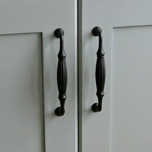 May include: Two matte black cabinet pulls with a vertical fluted design and curved ends, mounted onto light gray cabinet doors. The hardware features a central ribbed section that tapers toward the circular mounting points at the top and bottom.